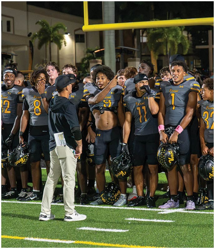 A group of people in black and yellow football uniforms stand in a line on the sidelines of a football field while a person in a black jacket and light pants stands facing them. Descripci n generada con IA