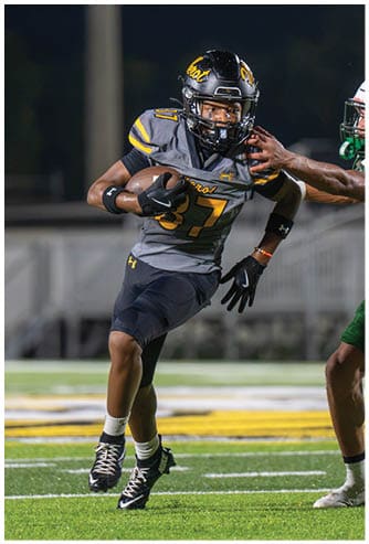 Two people in football uniforms are actively engaged in a game on a field, with one person running while holding the ball and the other reaching out to tackle. Descripci n generada con IA