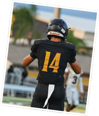 A person wearing a black football helmet with the brand name Riddell visible on the back is seen from behind outdoors with a blurred background of trees and buildings under a cloudy sky. Descripci n generada con IA