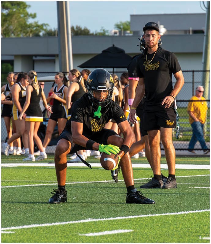 A person wearing a black helmet and sports gear is crouched holding a football on a field while another person in black athletic clothing stands nearby with a group of people in cheerleading uniforms in the background. Descripci n generada con IA