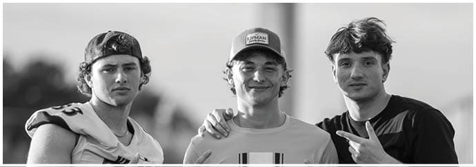 Three adults stand outdoors with one wearing a sports jersey and cap, another in a casual shirt and cap smiling with an arm around the first, and the third making a peace sign while wearing a dark shirt. Descripci n generada con IA