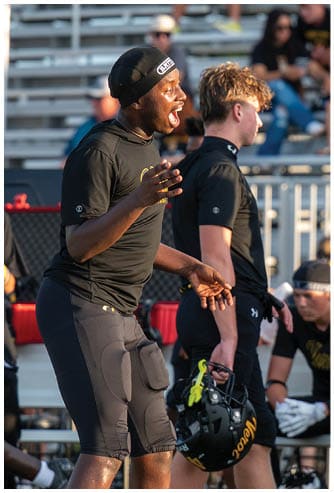A person wearing a black beanie and black shirt is shouting or cheering energetically in an outdoor setting with blurred bleachers and people in the background. Descripci n generada con IA