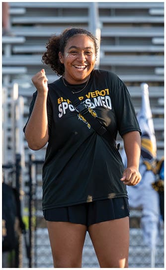 A person wearing a dark sports shirt and shorts is smiling and standing in front of bleachers while another person in a cheerleading uniform is blurred in the background. Descripci n generada con IA