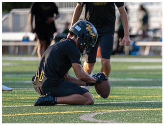 A person wearing a helmet and sports uniform is kneeling on a football field holding a football while another person stands nearby. Descripci n generada con IA
