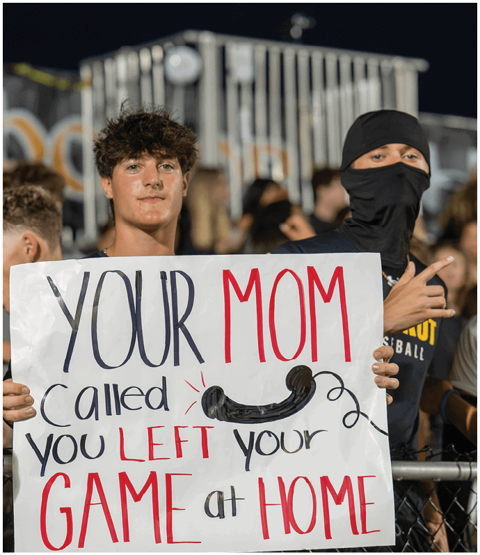 A person with curly hair holds a sign at a crowded event that reads “YOUR MOM called you LEFT your GAME at HOME" with some words in red and others in black. Descripci n generada con IA
