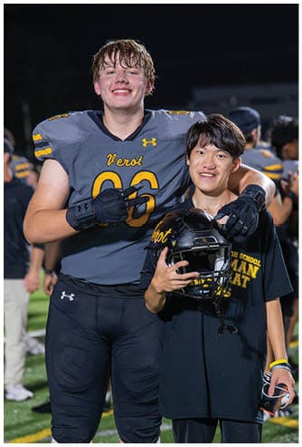 Two people wearing football uniforms smile while one holds a helmet and the other makes a peace sign with a gloved hand. Descripci n generada con IA