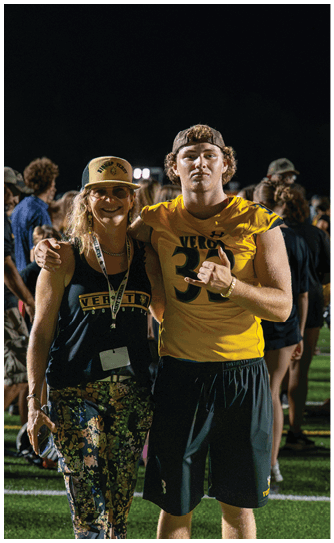Two people stand on a sports field at night with one person wearing a yellow sports jersey and the other wearing a hat and floral pants, while other people are visible in the background. Descripci n generada con IA
