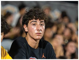 A person with curly hair and a black shirt is looking forward with a neutral expression against a blurred background of people. Descripci n generada con IA