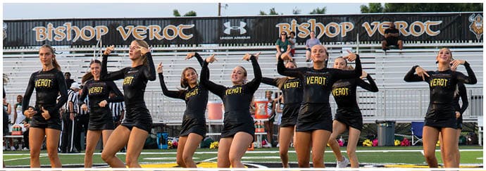 A group of people wearing black and gold uniforms are performing a synchronized cheer or dance routine in front of empty bleachers with a banner that reads Bishop Verot. Descripci n generada con IA