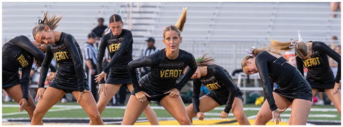 Adults in matching black athletic outfits with the word “VEROT" perform a synchronized dance routine on a sports field. Descripci n generada con IA