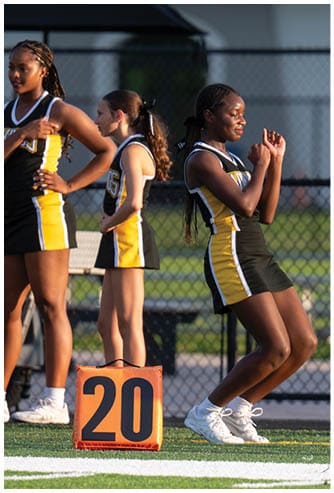 A person in a cheerleading uniform is standing on a sports field near an orange marker, appearing to be in mid-motion with a focused expression. Descripci n generada con IA