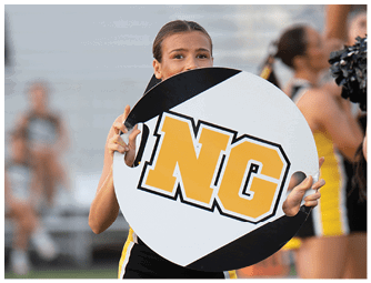 A person with pulled-back hair is holding a large circular sign with bold yellow and black letters while standing in an outdoor setting with blurred people in the background. Descripci n generada con IA