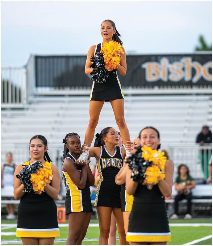 A person wearing a black cheerleading uniform with yellow and white stripes is standing on the shoulders of others while holding yellow and black pom-poms. Descripci n generada con IA