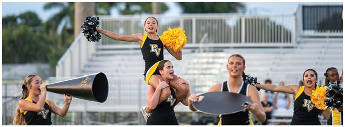 A group of cheerleaders in black and yellow uniforms perform a cheer routine on a sports field with one person lifted on another's shoulders holding pom-poms. Descripci n generada con IA