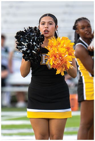 A person in a black and yellow cheerleading uniform holds black and yellow pom-poms while performing on a sports field. Descripci n generada con IA