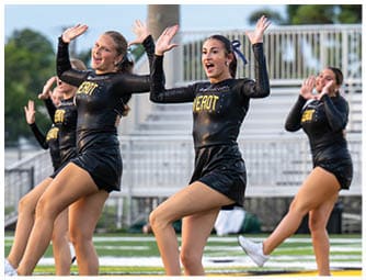 Adults in matching black cheerleading uniforms with the word “HEROT" perform a synchronized dance routine on a sports field. Descripci n generada con IA