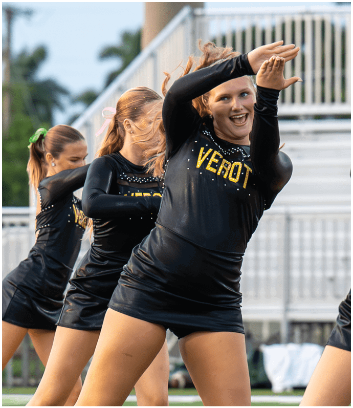 Adults in matching black and gold cheerleading uniforms perform a synchronized dance routine outdoors near bleachers. Descripci n generada con IA