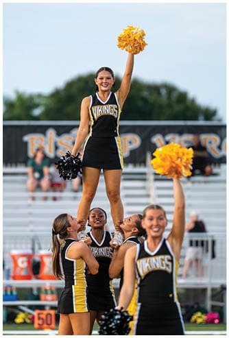A group of people in black and yellow cheerleading uniforms with “Vikings" written on them are performing a cheer stunt outdoors with one person lifted above the others holding yellow pom-poms. Descripci n generada con IA