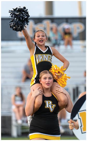 Two people in cheerleading uniforms with “Vikings" and "RV" logos perform a cheer stunt with one person sitting on the other's shoulders holding black and yellow pom-poms. Descripci n generada con IA