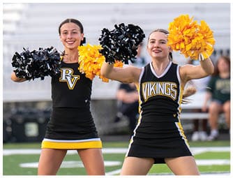 Two adults wearing black and yellow cheerleading uniforms are smiling and holding black and yellow pom-poms while performing on a sports field. Descripci n generada con IA