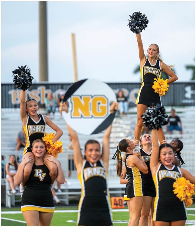 People in cheerleading uniforms are performing a routine in a stadium with one person holding a sign that says NG. Descripci n generada con IA