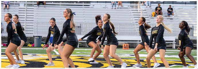 A group of people dressed in black athletic outfits are performing a synchronized dance or cheer routine on a sports field with empty bleachers in the background. Descripci n generada con IA