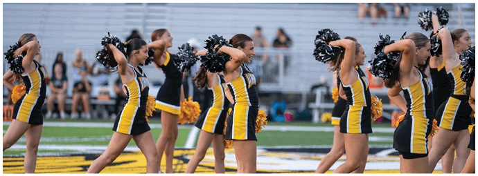 Adults in matching yellow and black cheerleading uniforms perform a synchronized routine on a sports field while holding black and yellow pom-poms. Descripci n generada con IA