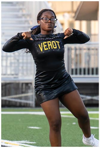 A person wearing glasses and a black cheerleading outfit with the word “VEROT" is performing a cheer pose on a sports field. Descripci n generada con IA