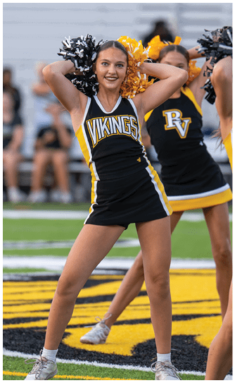 A person in a black and yellow Vikings cheerleading uniform is smiling and holding black and yellow pom-poms behind their head on a sports field. Descripci n generada con IA
