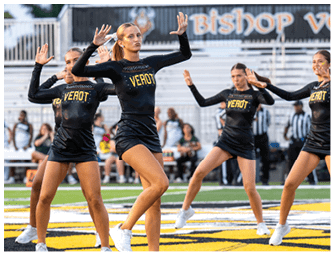 Adults in matching black athletic outfits with the word “VEROT" perform a synchronized dance routine on a sports field with bleachers and spectators in the background. Descripci n generada con IA