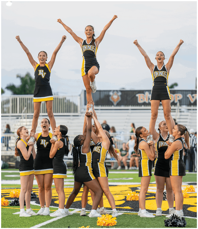 A group of cheerleaders in black and yellow uniforms perform a coordinated stunt on a sports field with some standing on the ground supporting others elevated in the air. Descripci n generada con IA