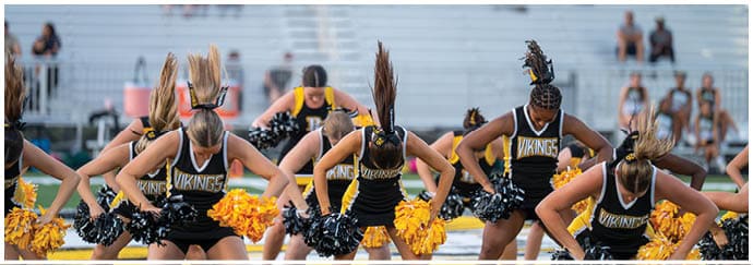A group of adults in black and yellow cheerleading uniforms with “Vikings" written on them perform a synchronized routine with pom-poms on a sports field with empty bleachers in the background. Descripci n generada con IA