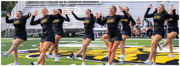 A group of adults dressed in black cheerleading uniforms with “DETROIT" written on them are performing a synchronized dance routine on a football field. Descripci n generada con IA