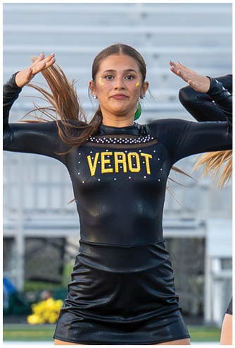 A person with long hair in a black shiny cheerleading outfit with the word “VEROT" in gold letters is performing a dance move with arms raised near the head on a sports field. Descripci n generada con IA