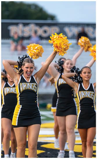 A group of adults in black and yellow cheerleading uniforms with “VIKINGS" on the front are performing with yellow pom-poms on a sports field. Descripci n generada con IA