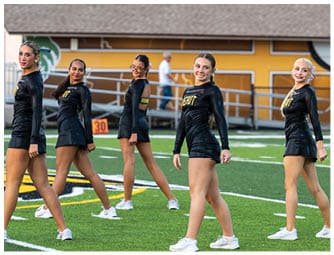 Five adults wearing black athletic outfits and white sneakers are standing in a line on a sports field with green turf and a yellow building in the background. Descripci n generada con IA