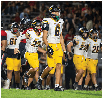 Two people wearing football helmets and white and yellow jerseys with the word “VEROT" and numbers are on a field, with one standing in the foreground and the other slightly behind. Descripci n generada con IA
