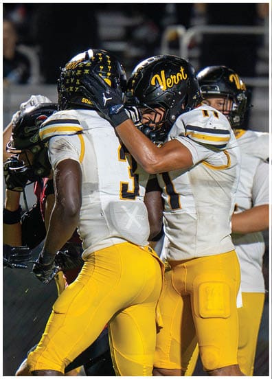 Two people in yellow football pants and white jerseys with black helmets are embracing on a football field during a game. Descripci n generada con IA