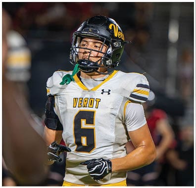 A person wearing a football helmet and a white jersey with the number 6 and the word “VEROT" stands on a field with a focused expression. Descripci n generada con IA