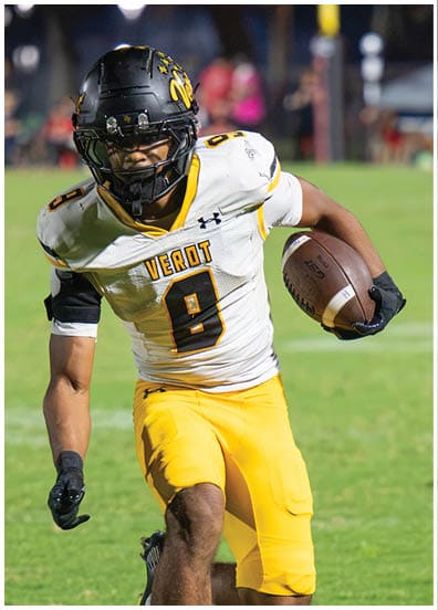 A person in a white and yellow football uniform is running with a football while another person in a red and black uniform is chasing them on a grassy field. Descripci n generada con IA