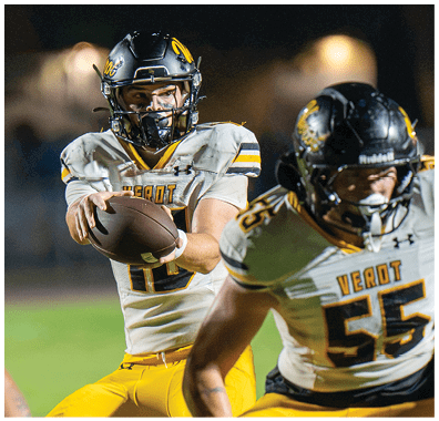A person wearing a football helmet and uniform is holding a football, preparing to throw it during a game. Descripci n generada con IA