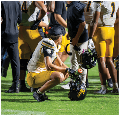 A person in a football uniform is squatting on the field holding a helmet and water bottle while other players and staff stand nearby. Descripci n generada con IA