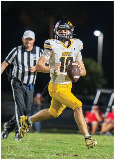 A person wearing a football uniform with the number 10 is running on a grassy field while holding a football, with a referee and blurred spectators in the background. Descripci n generada con IA