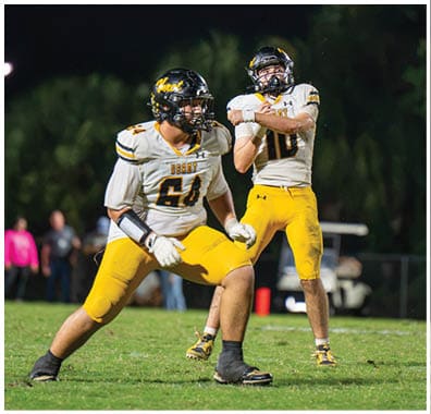 Two people wearing football uniforms and helmets are on a grassy field, one in a ready stance and the other preparing to throw a football. Descripci n generada con IA