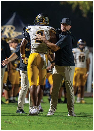 A person wearing a cap and headset is placing a hand on the back of a football player in yellow pants and a white jersey during a nighttime game. Descripci n generada con IA
