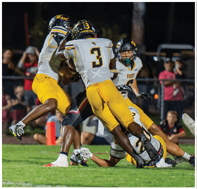 Three football players in yellow and white uniforms are tackling a player in a black uniform during a nighttime game. Descripci n generada con IA
