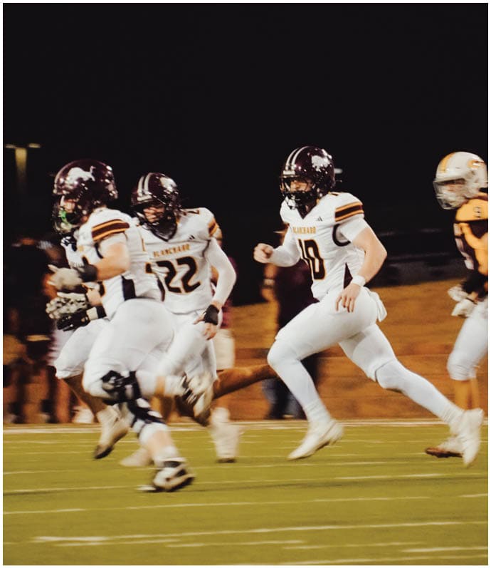 Three football players in blue and white uniforms running on a field. Description generated by AI
