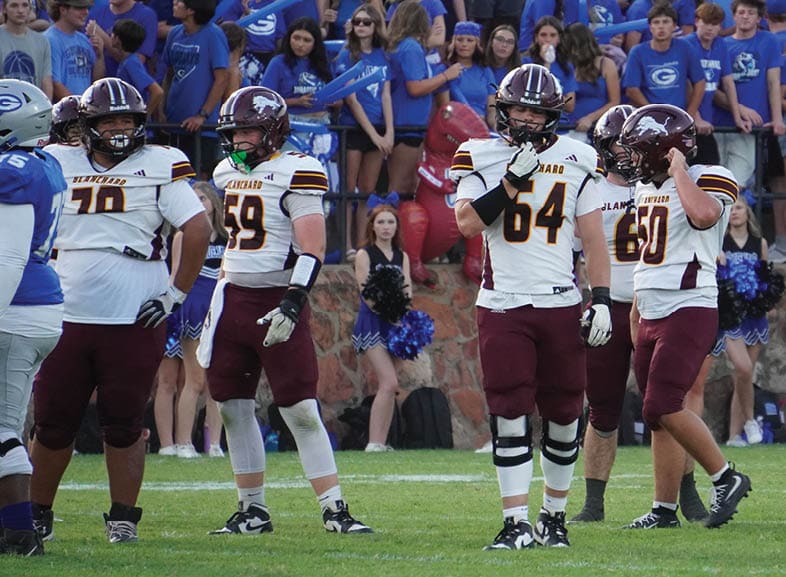 Four people wearing black football uniforms with “VISTA" on the front are standing together in a stadium with a crowd in the background, two of them holding helmets raised above their heads. Description generated by AI