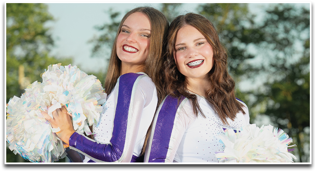 Two smiling adults with long hair and braces pose closely together outdoors with trees in the background. Description generated by AI
