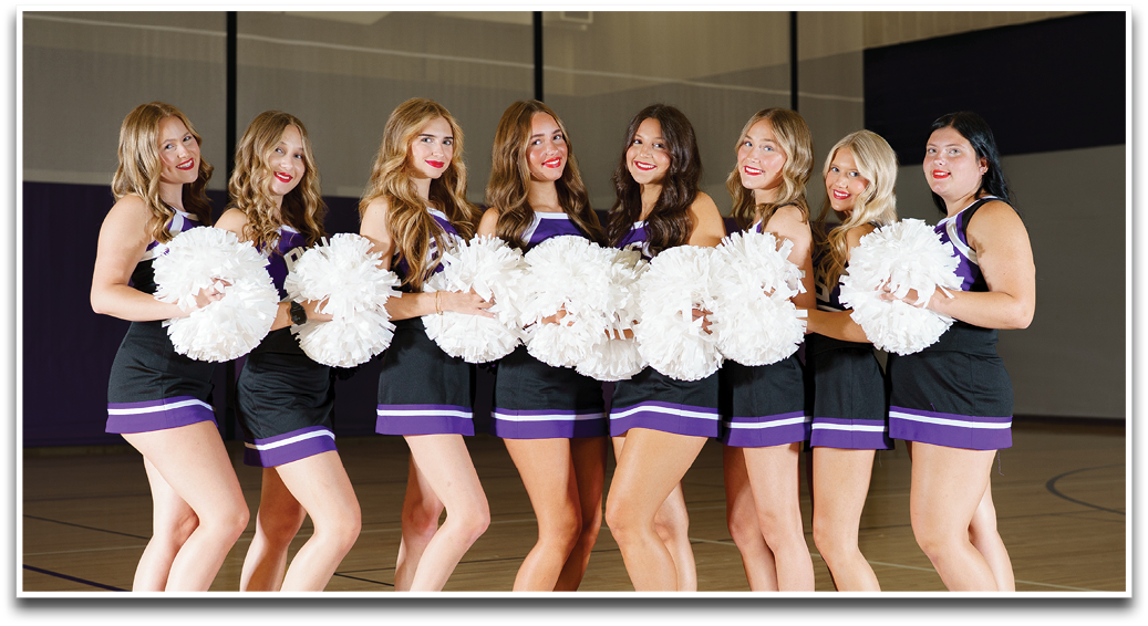A group of people wearing matching purple and white cheerleading uniforms pose together on a sports field with smiles. Description generated by AI